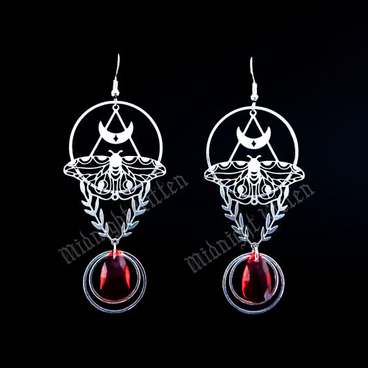 Pair of earrings with an intricate moth and laurel design and red stones on a black background