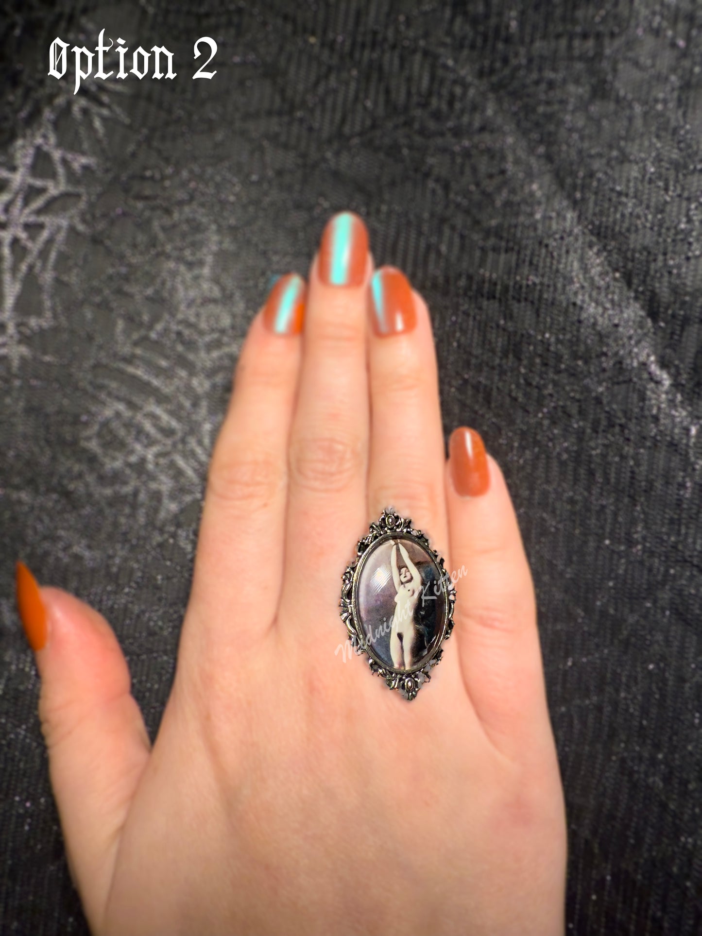 A silver ring with the decorative setting of a framed photograph on a hand against a textured black background. Option 2 contains a Victorian or Edwardian image of a woman bound by her hands which are tied above her head.